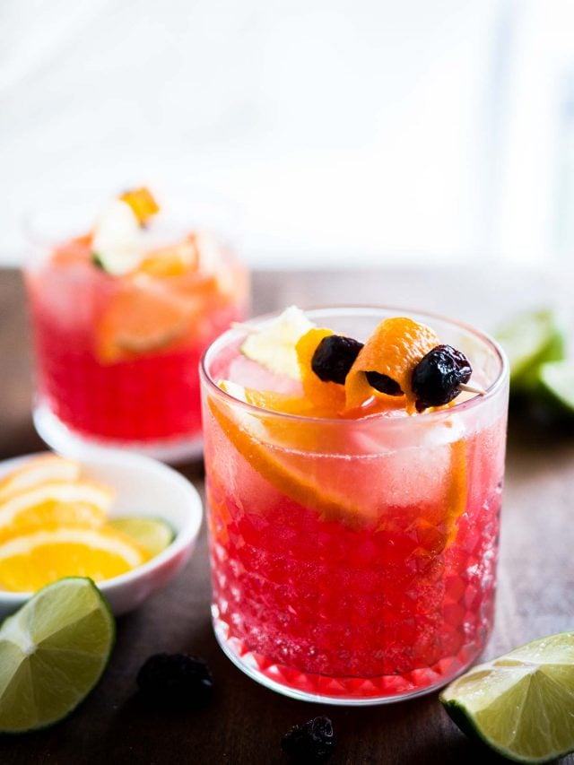 Close-up of two glasses of orange cranberry Moscow mule with fruit skewers. There\'s a small white bowl of lime and orange wedges as well as halved limes next to it.