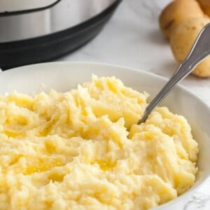 A white bowl of mashed potatoes with a spoon in it on a marble surface. There are potatoes and an instant pot in the background.